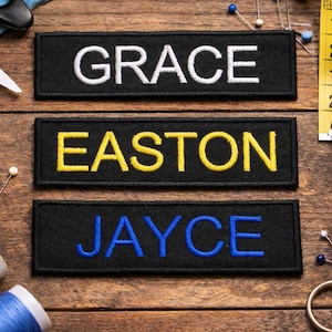 Puede incluir: Tres parches negros bordados con los nombres "GRACE", "EASTON" y "JAYCE" en hilo blanco, amarillo y azul. Suministros de costura, incluyendo tijeras, alfileres y carretes de hilo, están dispuestos sobre una superficie de madera.