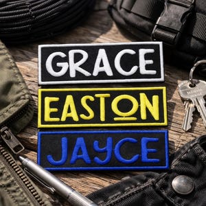 May include: Three embroidered name patches: "GRACE" in white on black, "EASTON" in yellow on black, and "JAYCE" in blue on black. The patches are rectangular and lie on a wooden surface, with a pen, keys, and clothing in the background.