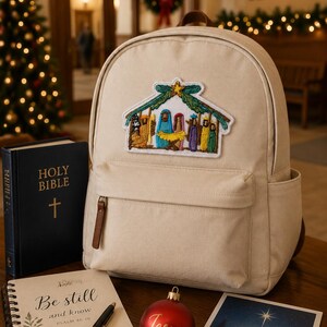 May include: A beige canvas backpack featuring an embroidered nativity scene patch. The backpack is displayed alongside a black Holy Bible, a spiral-bound notebook with the text Be still and know, a red ornament with the word Jesus, and a card.