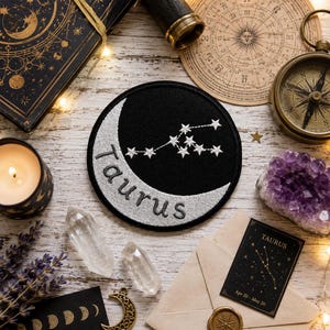 May include: A circular black embroidered patch featuring a silver crescent moon and the white stitched constellation of Taurus. The word Taurus is embroidered in silver thread along the curve of the moon. The patch is surrounded by various items.