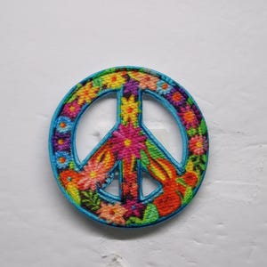 K&ouml;nnte beinhalten: Gestickter Peace-Zeichen-Aufn&auml;her mit einem lebendigen Blumenmuster. Der Aufn&auml;her hat eine blaue Umrandung, ein zentrales Friedenssymbol und ist mit bunten Blumen und orangefarbenen Hasen gef&uuml;llt. Ein fr&ouml;hliches Accessoire im Retro-Stil.
