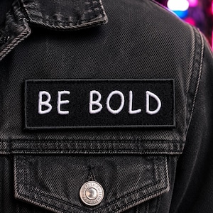 May include: A black rectangular patch with the words "BE BOLD" embroidered in white. The patch is affixed to a black denim jacket. The jacket has a buttoned pocket and a silver button.