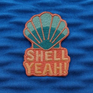 May include: Embroidered patch featuring a seashell design in teal and coral colors, with the words "SHELL YEAH!" in coral. The patch is likely for clothing or accessories, adding a fun, beachy vibe.