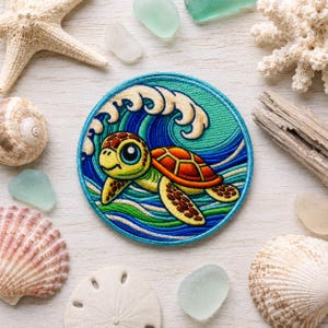 Op de afbeelding: Een ronde geborduurde patch met een cartoonzeeschildpad met een gele kop en oranje schild die door gestileerde blauwe en groene oceaangolven met witte schuimkoppen zwemt, omringd door diverse schelpen en stukjes zeeglas.
