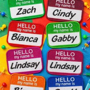 May include: A collection of colorful name tag patches with the text "HELLO my name is" and various names like Zach, Cindy, Bianca, Gabby, Lindsay, and Scott. The patches are in different colors, including green, burgundy, red, pink, orange, and blue.