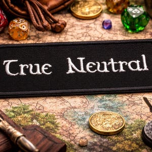 Puede incluir: Un parche negro bordado con las palabras "True Neutral" en blanco, rodeado de varios dados, monedas y una figura en miniatura. Los objetos están dispuestos sobre una superficie similar a un mapa, lo que sugiere un tema de juego de mesa.