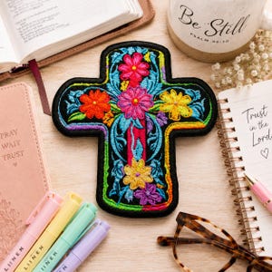 May include: A cross-shaped embroidered patch featuring a black background with vibrant, multi-colored floral patterns in shades of pink, orange, yellow, and blue, accented by intricate green leaf designs and a rainbow-colored border trim.