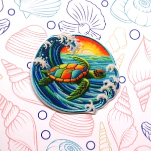 Peut inclure: &Eacute;cusson brod&eacute; repr&eacute;sentant une tortue de mer nageant devant une vague avec un coucher de soleil. La carapace de la tortue est color&eacute;e, avec des nuances d'orange, de vert et de bleu. L'&eacute;cusson rond a une bordure blanche, sur un fond d'illustrations de coquillages.