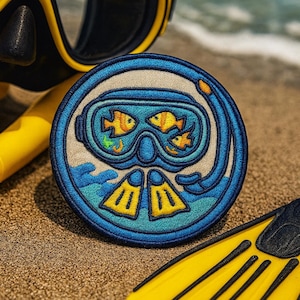 May include: A round, embroidered patch featuring a scuba mask with three yellow fish inside. The patch is blue and white, with yellow fins at the bottom. A yellow snorkel and fins are in the background, on a sandy beach.
