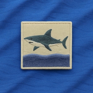 Peut inclure: Écusson brodé avec un motif de requin. Le requin est représenté dans des tons de bleu et de blanc, sur un fond beige. En dessous du requin, un motif de vagues bleues. L'écusson carré a une bordure blanche, sur un tissu bleu.