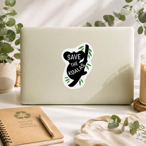 May include: A white sticker on a laptop features a black koala silhouette clinging to a branch, with the text "Save the Koalas" in white. Green leaves accent the design. Other items include a notebook, pen, and a tote bag.