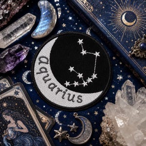 以下が含まれることがあります： 黒い背景に白い三日月と水瓶座の星座が刺繍された円形のワッペン。月の曲線に沿って白い小文字で「aquarius」という文字が刺繍されています。ワッペンには黒い縁取りが施されています。
