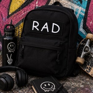 May include: A black backpack featuring a rectangular patch with the white text RAD, accompanied by a black water bottle with smiley face and skeleton hand stickers, black over-ear headphones, and a black notebook with a white melting smiley face.