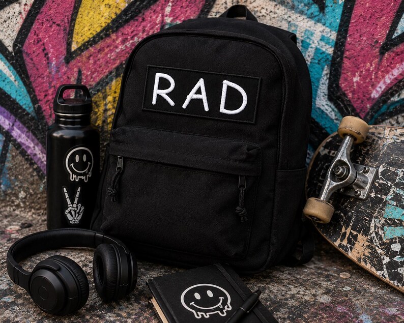 May include: A black backpack featuring a rectangular patch with the white text RAD, accompanied by a black water bottle with smiley face and skeleton hand stickers, black over-ear headphones, and a black notebook with a white melting smiley face.