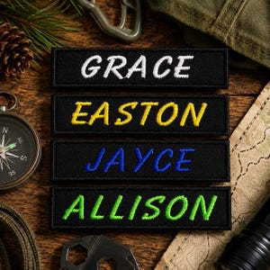 Puede incluir: Cuatro parches rectangulares negros bordados con nombres en diferentes colores de hilo. El parche superior muestra el nombre GRACE en blanco, el segundo muestra EASTON en amarillo, el tercero presenta JAYCE en azul y el parche inferior muestra ALLISON en verde lima.