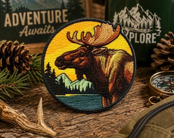 May include: A circular embroidered patch featuring a brown moose in profile against a yellow sunset sky, green mountains, and a dark blue lake. The patch has a black stitched border and depicts a detailed wilderness scene with pine trees.