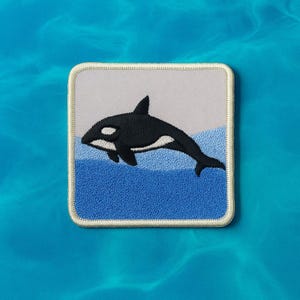 May include: Embroidered patch featuring a black and white orca whale leaping from blue water. The patch has a light gray background above the water line and a white border. The orca is a popular marine mammal.