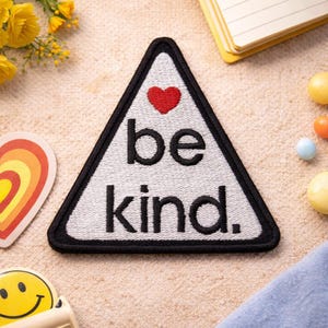 Puede incluir: Un parche triangular con fondo blanco y borde negro. El parche presenta las palabras "be kind" en negro, con un corazón rojo encima de la palabra "be". El parche está rodeado de otros objetos pequeños.