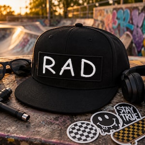 May include: A black baseball cap with a rectangular patch featuring the white embroidered text RAD. The cap is surrounded by black sunglasses, a black T-shaped tool, black headphones, and three stickers including a smiley face and a skateboard.