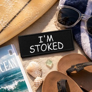 May include: A rectangular black embroidered patch featuring the white text I'M STOKED in a casual font. The patch is positioned on a sandy surface alongside a surfboard, clear sunglasses, a blue and white striped towel, seashells, and a magazine.
