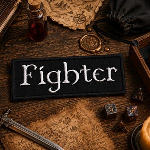 以下が含まれることがあります： 黒い布製の長方形のワッペンで、白い糸でFighterという文字が太くはっきりとしたフォントで刺繍されています。ワッペンにはステッチが施された縁があり、平らな長方形の布製品として、読みやすい文字が特徴です。