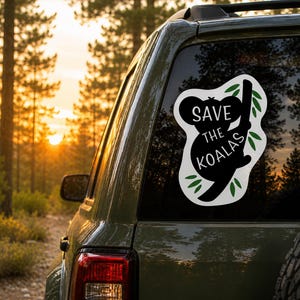 May include: A black and white sticker on a car window features a koala silhouette clinging to a branch, with the text "SAVE THE KOALAS." Green leaves surround the koala. The background shows a forest at sunset.