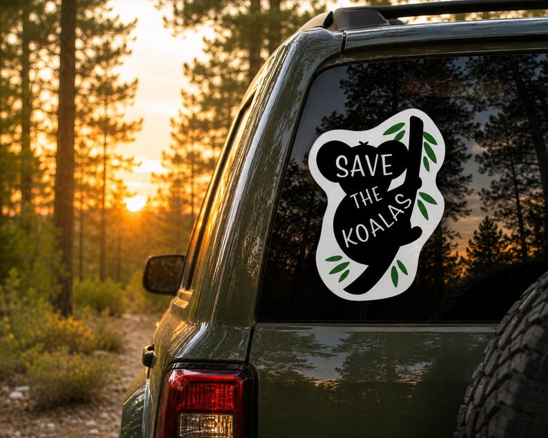 May include: A black and white sticker on a car window features a koala silhouette clinging to a branch, with the text "SAVE THE KOALAS." Green leaves surround the koala. The background shows a forest at sunset.