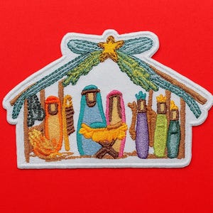 May include: An embroidered patch featuring a nativity scene inside a stable structure. The design includes stylised figures in orange, blue, pink, purple, green, and teal, with a yellow star at the top. The patch has a white border and white background.