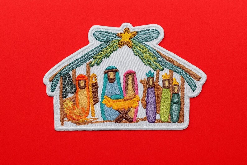 May include: An embroidered patch featuring a nativity scene inside a stable structure. The design includes stylised figures in orange, blue, pink, purple, green, and teal, with a yellow star at the top. The patch has a white border and white background.