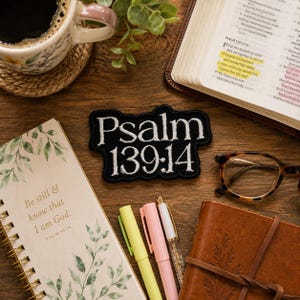 May include: A black embroidered patch with white text reading Psalm 139:14, positioned among a coffee mug, an open Bible, a spiral-bound notebook with the text Be still & know that I am God, a brown leather journal, and three pens.