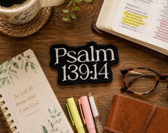 May include: A black embroidered patch with white text reading Psalm 139:14, positioned among a coffee mug, an open Bible, a spiral-bound notebook with the text Be still & know that I am God, a brown leather journal, and three pens.