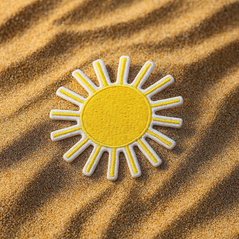 May include: A yellow sun patch with white trim, resting on a sandy surface. The sun design features a bright yellow center and white rays, creating a cheerful and summery aesthetic. The patch is likely for clothing or accessories.