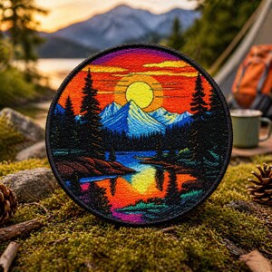 May include: Circular embroidered patch depicting a sunset over a mountain lake. The design shows a yellow sun, blue mountains, and colourful reflections in the water. The patch has a black border and is likely aimed at outdoor enthusiasts.