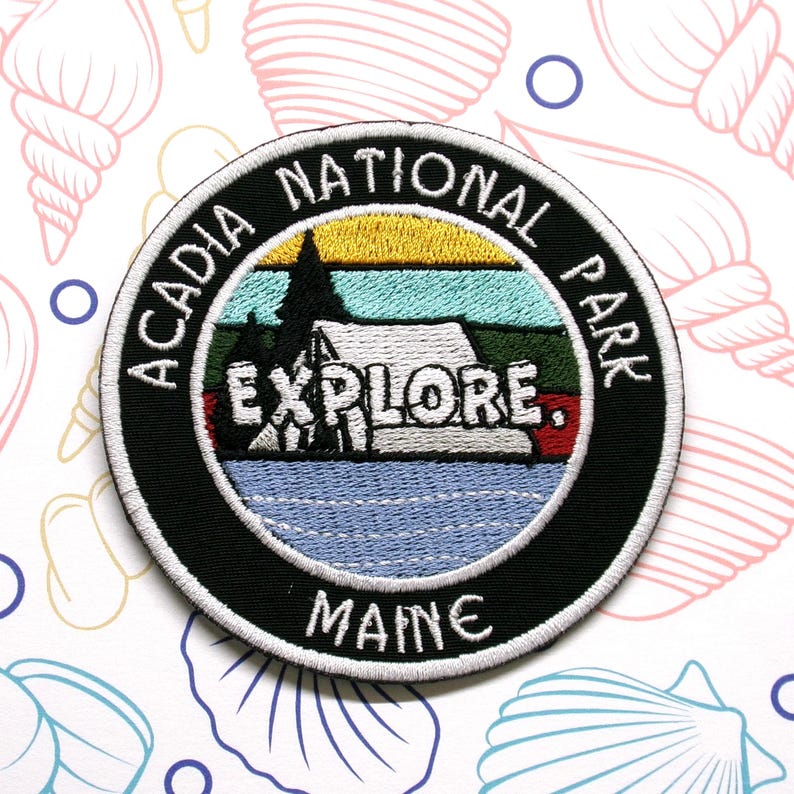 Może przedstawiać: Okrągła, czarno-biała naszywka haftowana z tekstem "Acadia National Park" i "Maine" w kolorze białym. Środek naszywki przedstawia kolorową ilustrację namiotu, drzew i jeziora z napisem "Explore" w kolorze czerwonym.