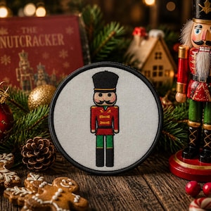 Könnte beinhalten: Ein kreisförmiger Stickaufnäher mit einer Nussknacker-Figur, die einen schwarzen Hut, eine rote Jacke und eine grüne Hose auf weißem Grund trägt. Der Aufnäher hat einen dicken schwarzen Rand und wird als eigenständiger Dekorationsartikel gezeigt.