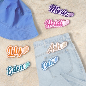 Peut inclure: Un chapeau de pêcheur bleu clair et des écussons brodés avec des noms sur un jean en denim délavé. Les écussons présentent des noms tels que "Marie", "Heidi", "Lily", "Eden", "Ash" et "Cali" en différentes couleurs avec des accents de coquillages.