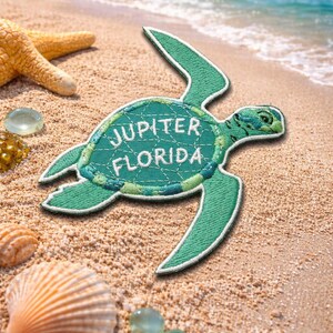 Pu&ograve; includere: Toppa ricamata turchese a forma di tartaruga marina, con la scritta "JUPITER FLORIDA" in bianco. La toppa &egrave; su una spiaggia sabbiosa con conchiglie, una stella marina e il mare sullo sfondo.