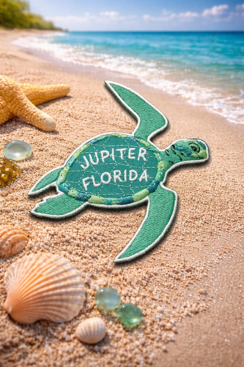 Pu&ograve; includere: Toppa ricamata turchese a forma di tartaruga marina, con la scritta "JUPITER FLORIDA" in bianco. La toppa &egrave; su una spiaggia sabbiosa con conchiglie, una stella marina e il mare sullo sfondo.