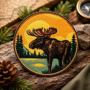 May include: Circular embroidered patch depicting a moose by a lake with a mountain backdrop. The design features a yellow and orange sunset, green trees, and a brown moose. The patch has a black border, suitable for outdoor enthusiasts or nature lovers.