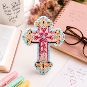 Könnte beinhalten: Ein bestickter Aufnäher in Kreuzform mit komplexen, floral inspirierten Mustern in Rosa, Gold und Hellblau. Das Kreuz hat einen dekorativen, gewellten Rand, der mit hellblauem Garn umrandet ist, sowie ein cremefarbenes Zentrum und symmetrische Details.