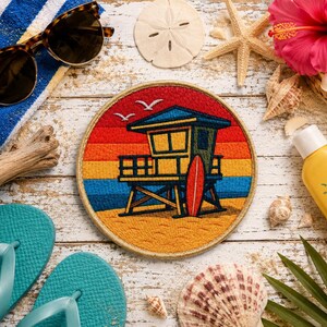 May include: A circular embroidered patch featuring a lifeguard tower on a beach with a red surfboard, set against a sunset background of red, orange, yellow, and blue horizontal stripes, with two white birds flying in the sky above the tower.