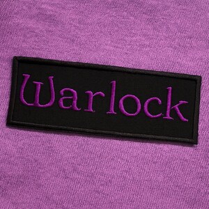 K&ouml;nnte beinhalten: Ein rechteckiger, schwarz bestickter Aufn&auml;her mit dem Wort Warlock in violettem Garn, der auf einen strukturierten, hellvioletten Stoffgrund gen&auml;ht ist. Der Aufn&auml;her hat einen sauberen schwarzen Rand und ist horizontal auf der violetten Textiloberfl&auml;che zentriert.