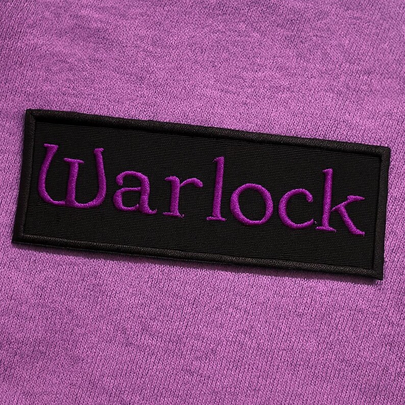 K&ouml;nnte beinhalten: Ein rechteckiger, schwarz bestickter Aufn&auml;her mit dem Wort Warlock in violettem Garn, der auf einen strukturierten, hellvioletten Stoffgrund gen&auml;ht ist. Der Aufn&auml;her hat einen sauberen schwarzen Rand und ist horizontal auf der violetten Textiloberfl&auml;che zentriert.