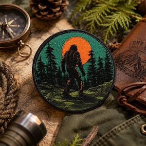 Puede incluir: Parche bordado redondo con la silueta de un Bigfoot frente a una puesta de sol. El parche tiene un borde verde y negro, con un fondo verde y un sol naranja. El diseño incluye pinos y un primer plano herboso.