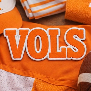 Pode incluir: Uma variedade de artigos de fã dos Tennessee Volunteers em laranja e branco, incluindo um boné de beisebol, pompons, uma bola de futebol americano e uma toalha. A palavra "VOLS" é exibida em laranja.