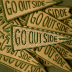Könnte beinhalten: Eine Ansammlung von bestickten Wimpel-Patches mit dem grünen Schriftzug "GO OUT SIDE". Die Patches sind beige mit grünen Rändern und einem grünen Pfeildesign. Sie liegen auf einem grünen Hintergrund.