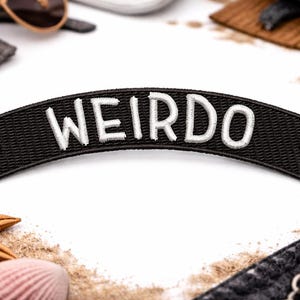 Może przedstawiać: Czarna naszywka z białym napisem "WEIRDO" w zakrzywionym kształcie banera. Obraz zawiera rozgwiazdę, muszlę i akcesoria na białym tle.