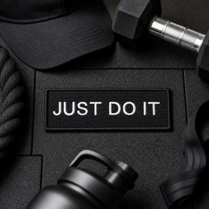 Może przedstawiać: Czarnobiała martwa natura o tematyce fitness. Zawiera czarną czapkę z daszkiem, czarną naszywkę z napisem "JUST DO IT", czarną hantle, czarną butelkę na wodę i czarną taśmę oporową. Przedmioty są ułożone na teksturowanej czarnej powierzchni.