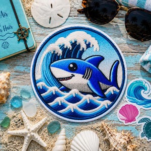 以下が含まれることがあります： 青い海の波の中を泳ぐ漫画のサメが描かれた円形の刺繍ワッペン。サメは青と白で、親しみやすい表情をしています。ワッペンには白い縁取りがあり、周囲には貝殻、ヒトデ、ステッカーが配置されています。