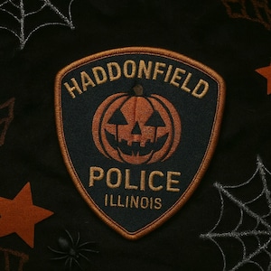 May include: A Halloween-themed patch with a pumpkin design inside a shield shape. The patch reads "HADDONFIELD POLICE ILLINOIS" in gold lettering. The background is black with spiderwebs and stars.
